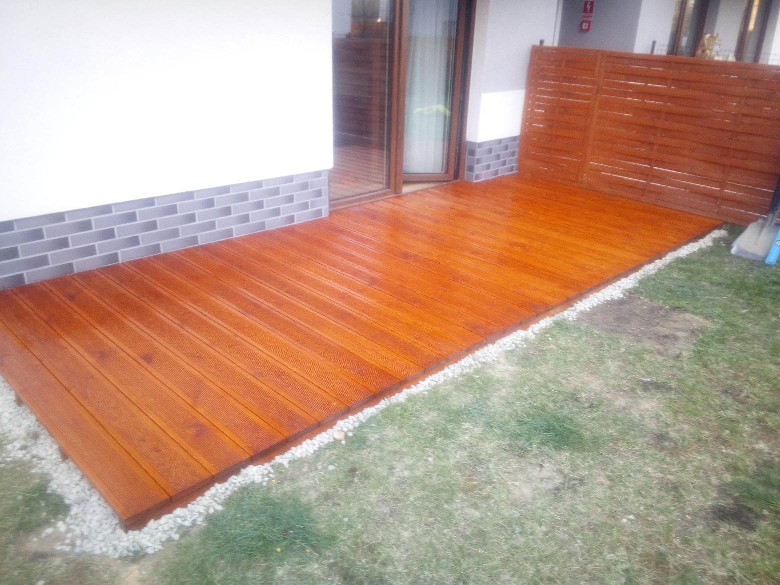 Świeżo zaimpregnowany, drewniany taras z desek ryflowanych, obrzeża wysypane drobnym, białym kamieniem, przylegający do ściany domu wykończonej szarą cegłą klinkierową i drewnianego ażurowego...