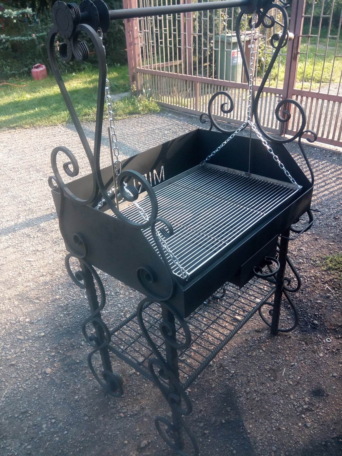 Kuty grill ogrodowy z ozdobnymi elementami, zawieszony na łańcuchach, z dolną półką na akcesoria, na tle metalowej bramy i zieleni.