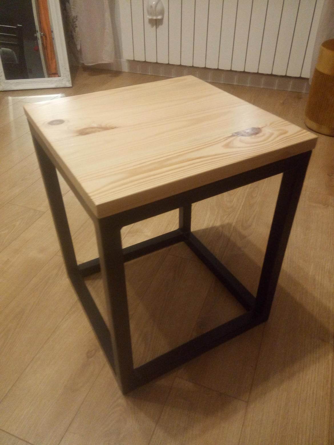 Drewniany taboret z blatem z jasnego drewna i czarną, metalową ramą, stojący na panelach podłogowych.