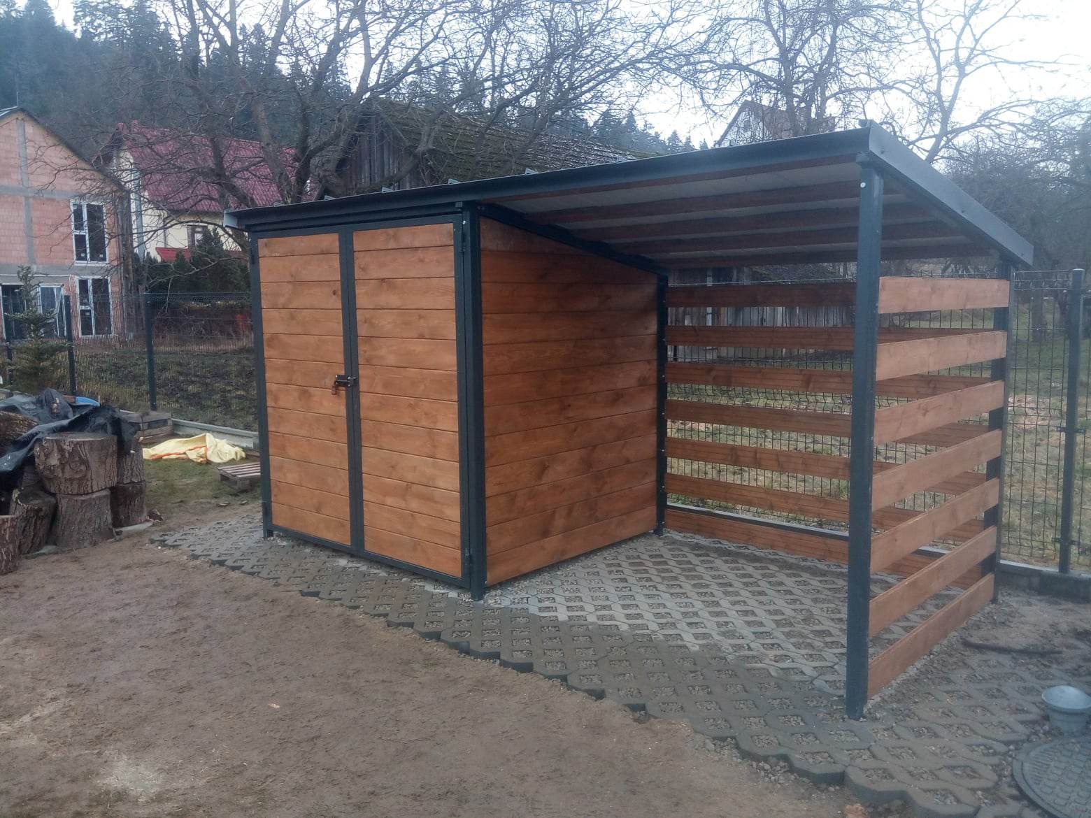 Drewniany schowek na narzędzia z zadaszeniem, obok ułożone drewno na tle domu w budowie i drzew bez liści.