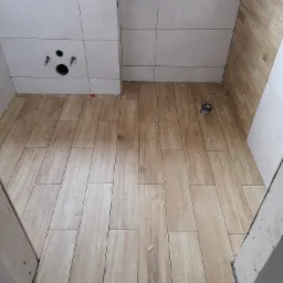 Ułożone płytki podłogowe imitujące drewno w łazience, widoczne otwory na armaturę i białe płytki ścienne, plastikowe krzyżyki dystansowe między płytkami.