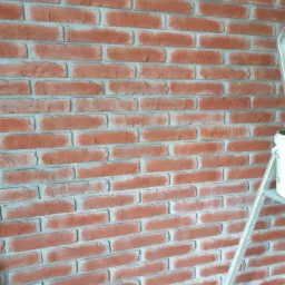 Ściana wykończona czerwoną cegłą dekoracyjną, widoczny fragment drabiny malarskiej i akcesoria malarskie na podłodze.