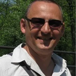 Portret mężczyzny w okularach przeciwsłonecznych na tle zieleni drzew.