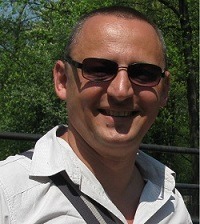 Portret mężczyzny w okularach przeciwsłonecznych na tle zieleni drzew.