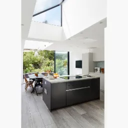 Nowoczesna kuchnia z dużym oknem dachowym i widokiem na zielony ogród, wyspa kuchenna z płytą indukcyjną i szufladami, stół jadalny z krzesłami w tle.
