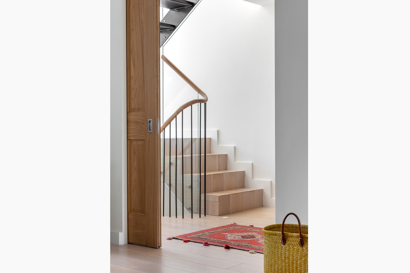 Otwarta, przesuwna dębowa drzwi ukazuje minimalistyczne schody z drewnianymi stopniami, czarną balustradą i białymi ścianami, uzupełnione orientalnym dywanikiem i słomkowym koszem.