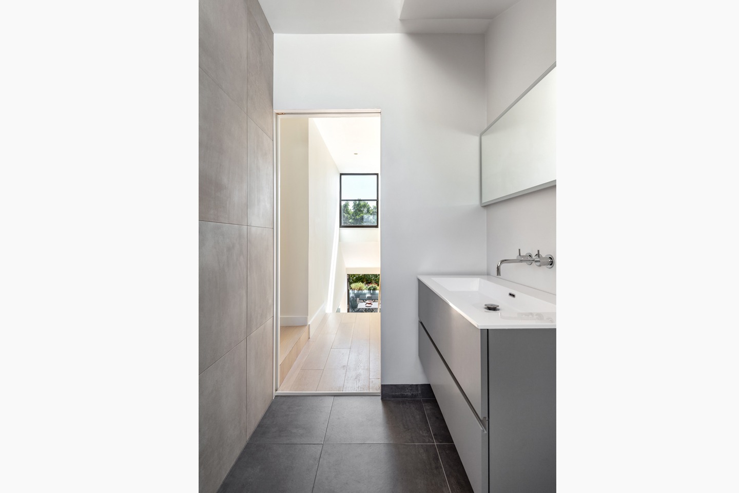 Nowoczesna łazienka z szarą zabudową umywalki, lustrem i widokiem na schody oraz ogród przez okno.