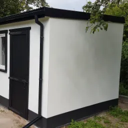 Wykończony, biały budynek gospodarczy z czarnymi elementami wykończeniowymi (dach, okna, drzwi, podstawa) w otoczeniu zieleni ogrodowej.