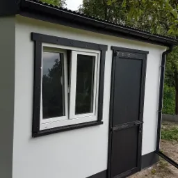 Ocieplony budynek gospodarczy w Zduńskiej Woli z czarną stolarką okienną i drzwiami, widoczny fragment rynny i korona drzewa.