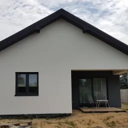 Nowoczesny dom jednorodzinny z jasną elewacją i ciemną stolarką okienną oraz dachem, widok z zewnątrz, w tle błękitne niebo z chmurami, widoczny żółty wąż do ocieplania.