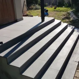 Wykończone schody wejściowe do domu, pokryte szarą, matową glazurą, z widocznymi spoinami i cieniem słupa, na tle zieleni ogrodu.