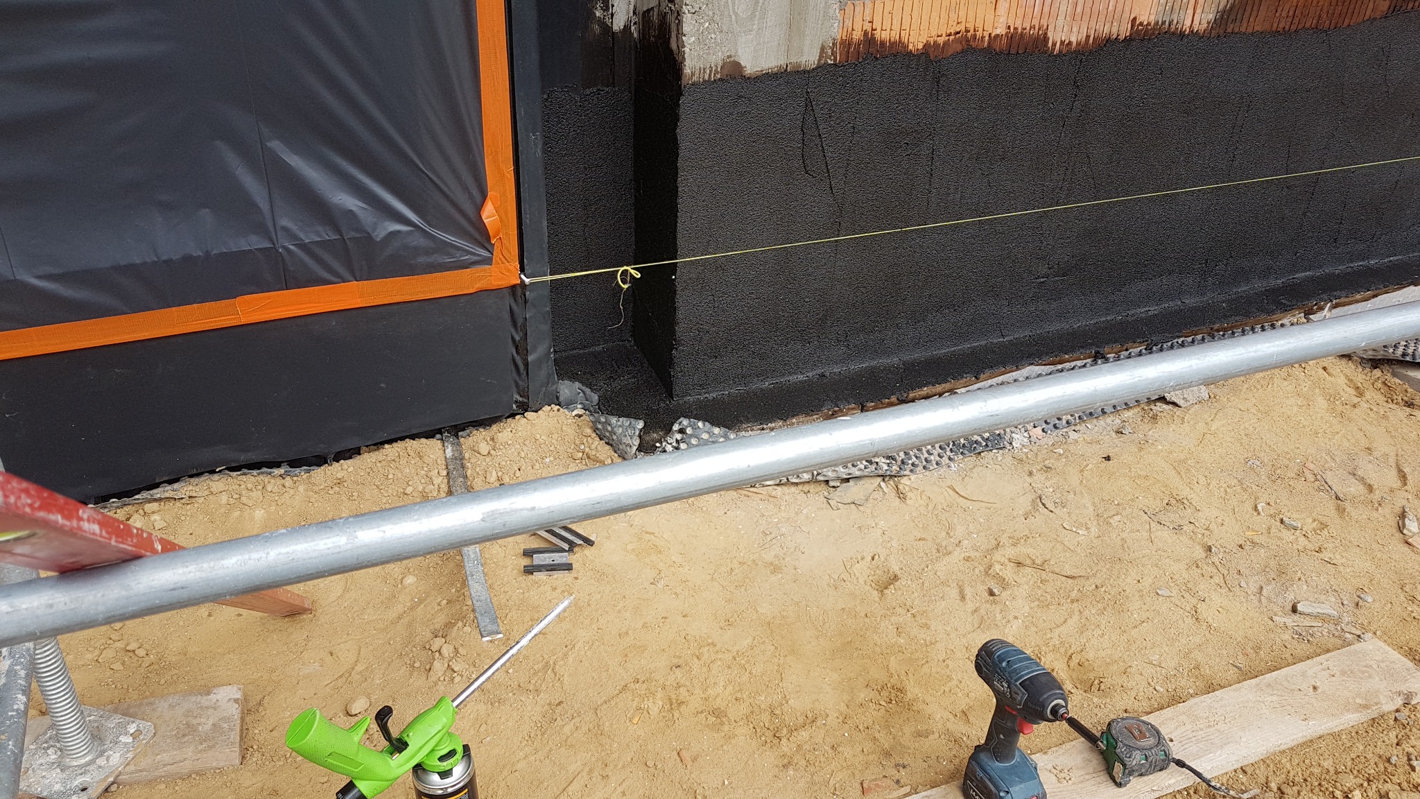 Izolacja fundamentów budynku w trakcie prac: czarna powłoka hydroizolacyjna, folia ochronna z pomarańczową taśmą, poziomica sznurkowa, narzędzia budowlane (wiertarka, pistolet do pianki)...