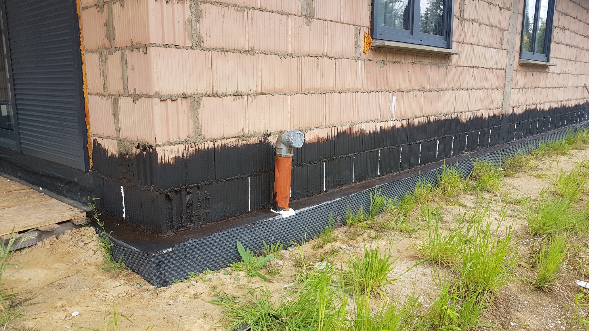 Świeżo zaizolowana ściana fundamentowa budynku z bloczków, pokryta czarną hydroizolacją i folią kubełkową, z wystającą rurą odpływową uszczelnioną pianką.