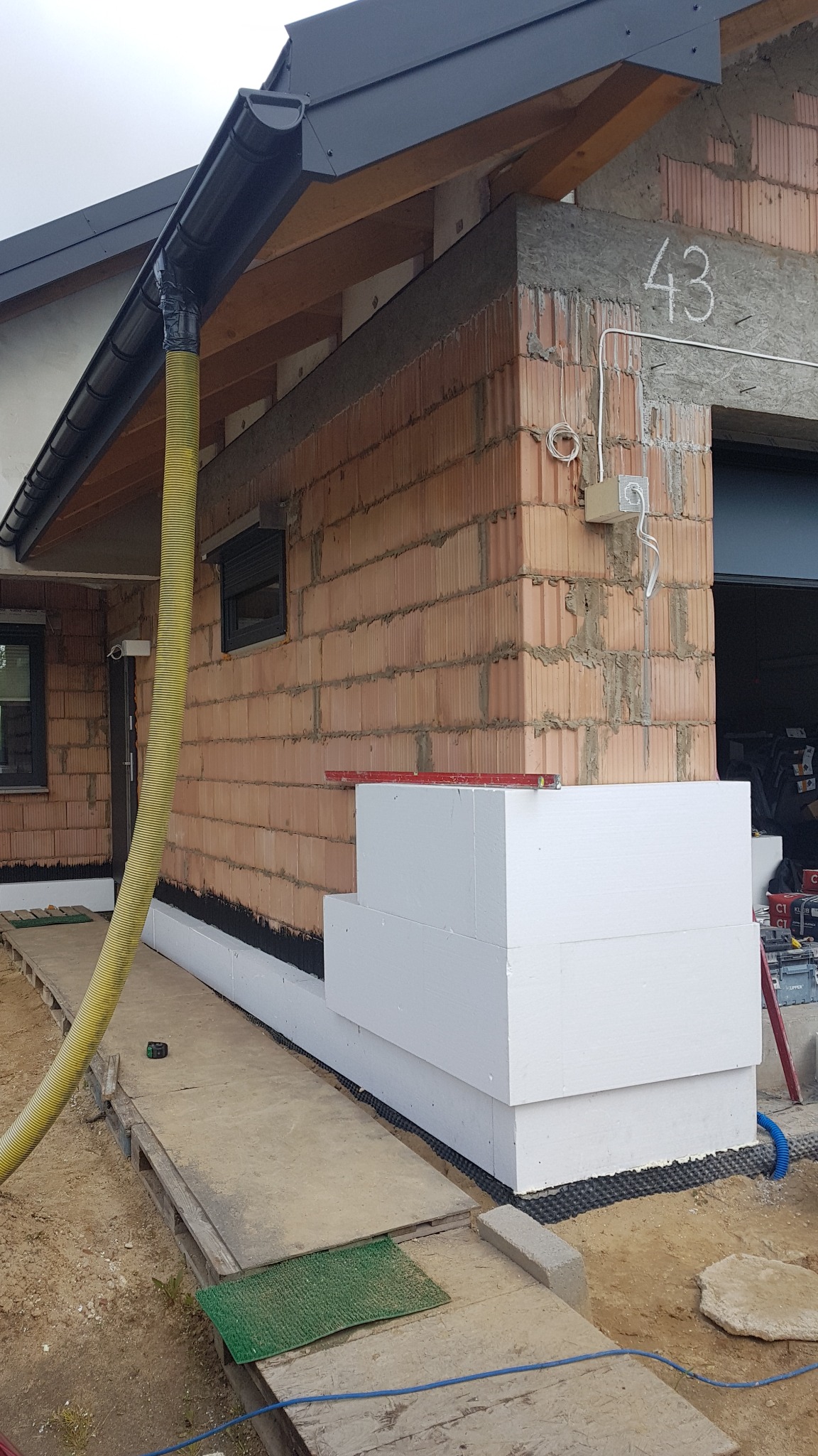 Fragment elewacji budynku w trakcie prac: widoczne bloczki z betonu komórkowego, warstwa izolacji ze styropianu, żółty wąż wentylacyjny oraz poziomica na styropianie.