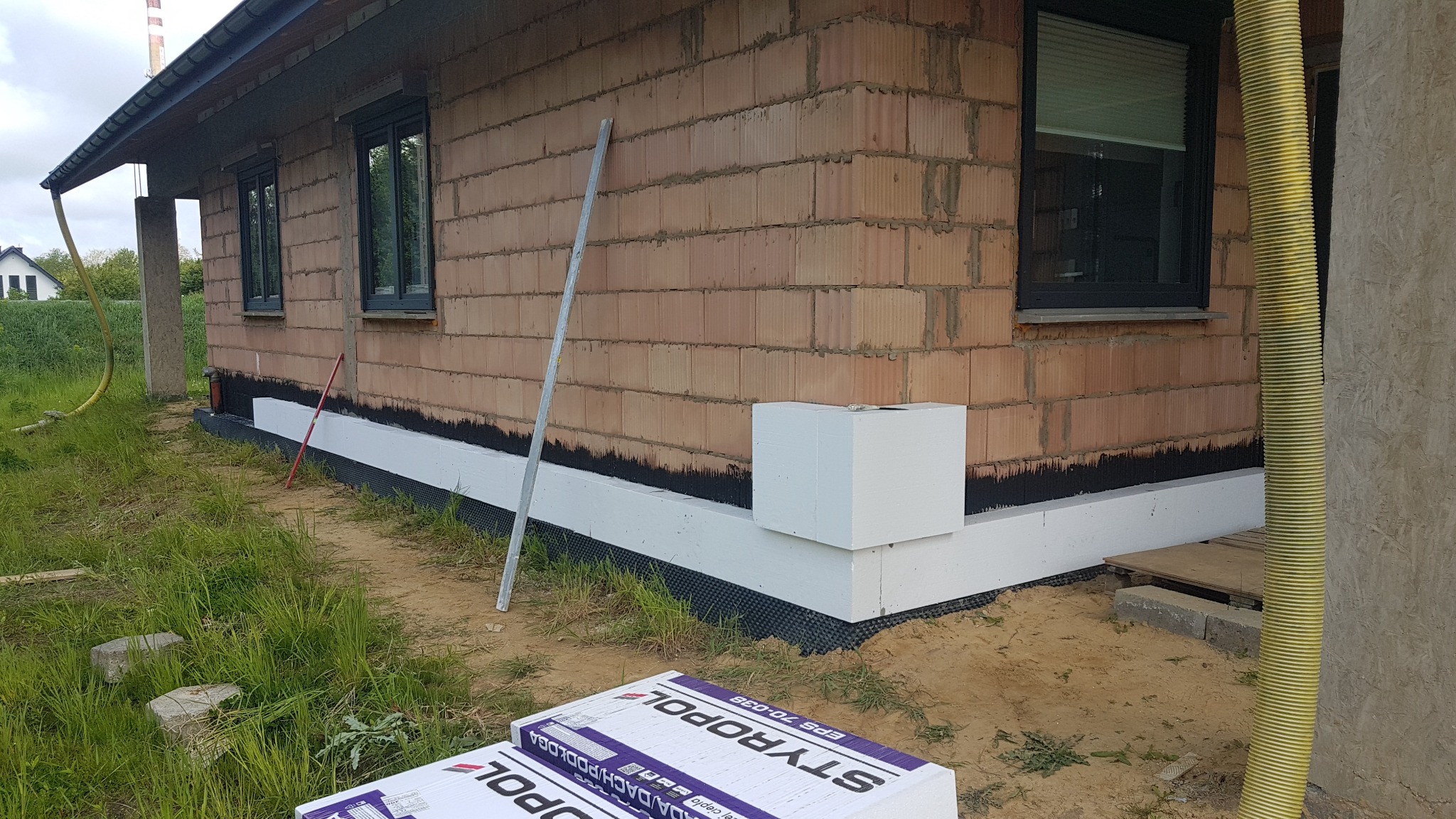 Fragment ściany budynku z widocznym ociepleniem fundamentu wykonanym ze styropianu, na pierwszym planie paczki styropianu Styropool, widoczne okna z czarną ramą i mur z czerwonej cegły dziurawki.