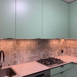 Dawid Ronek - Nowoczesna kuchnia z pistacjowymi szafkami, płytkami imitującymi kamień i oświetleniem LED pod szafkami wiszącymi, widoczny zlew z czarną baterią i płyta gazowa.