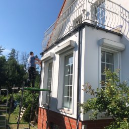 Dawid Ronek - Mężczyzna na rusztowaniu maluje elewację domu z balkonem i roletami okiennymi; widoczne narzędzia malarskie i drabina oparta o ścianę.