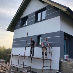Dawid Ronek - Dwóch mężczyzn na rusztowaniu maluje elewację domu w poziome pasy, szare i białe, z widocznymi oknami i fragmentem dachu.