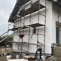 Dawid Ronek - Pracownik na rusztowaniu podczas nakładania białego tynku na elewację domu jednorodzinnego, widoczne puszki z farbą, styropian, okna zabezpieczone folią.