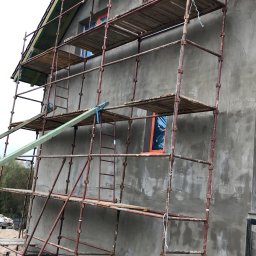 Dawid Ronek - Budynek w trakcie nakładania szarego tynku, z zamontowanym rusztowaniem i zabezpieczonymi oknami folią ochronną.