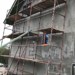 Budynek w trakcie nakładania szarego tynku, z zamontowanym rusztowaniem i zabezpieczonymi oknami folią ochronną.