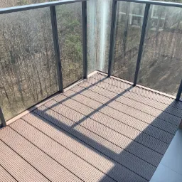 Nowoczesny balkon z szarą podłogą z desek kompozytowych i szklaną balustradą, widok na las w tle.