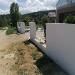 Nowoczesny, biały mur ogrodzeniowy w trakcie budowy, widoczny z perspektywy ziemi, z elementami szalunku i narzędziami, w tle dom z przeszklonym balkonem i zieleń.