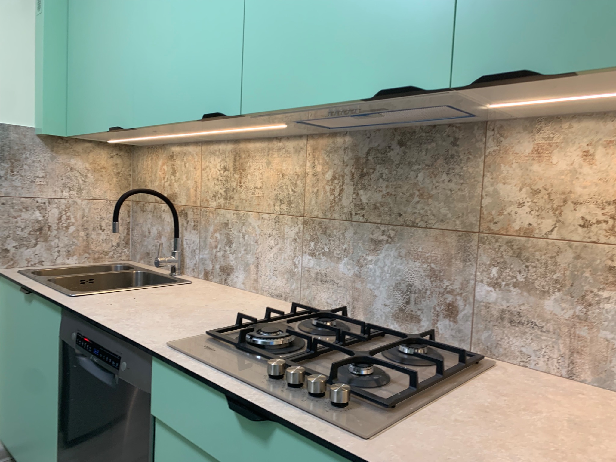 Nowoczesna kuchnia z miętowo-zielonymi szafkami, beżowym blatem, płytą gazową Whirlpool i zlewozmywakiem dwukomorowym pod czarną baterią.