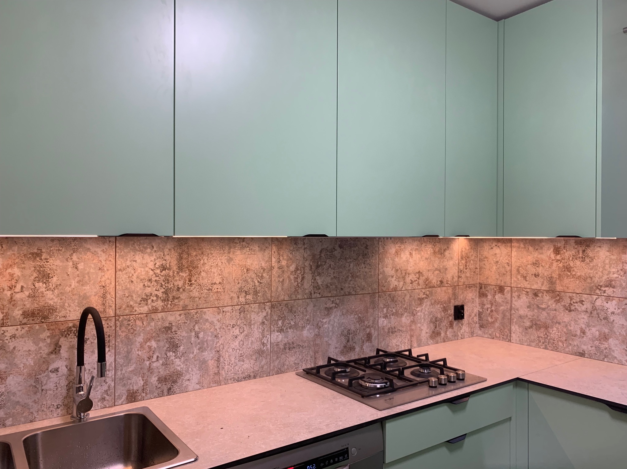 Nowoczesna kuchnia z pistacjowymi szafkami, płytkami imitującymi kamień i oświetleniem LED pod szafkami wiszącymi, widoczny zlew z czarną baterią i płyta gazowa.