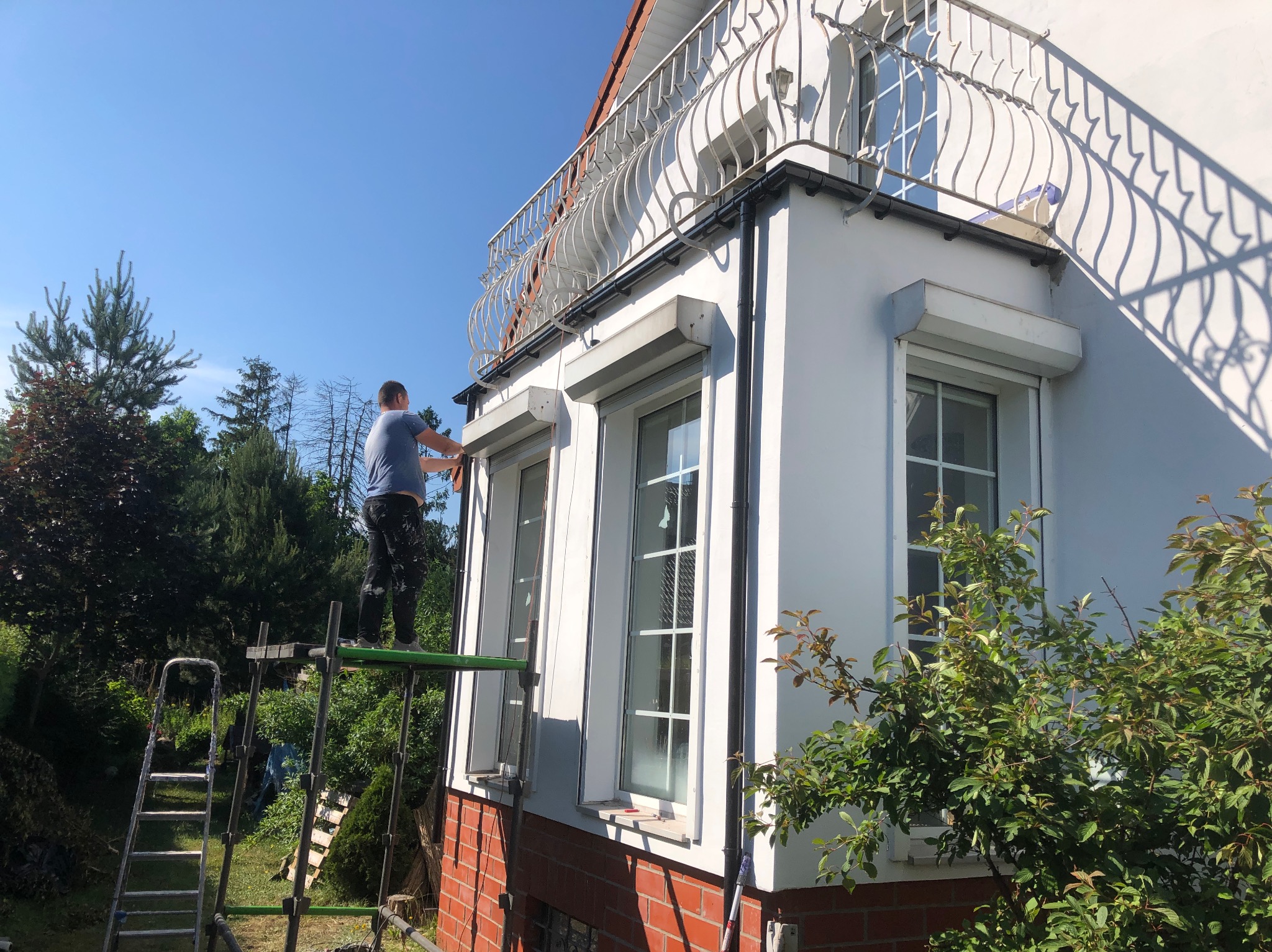 Mężczyzna na rusztowaniu maluje elewację domu z balkonem i roletami okiennymi; widoczne narzędzia malarskie i drabina oparta o ścianę.