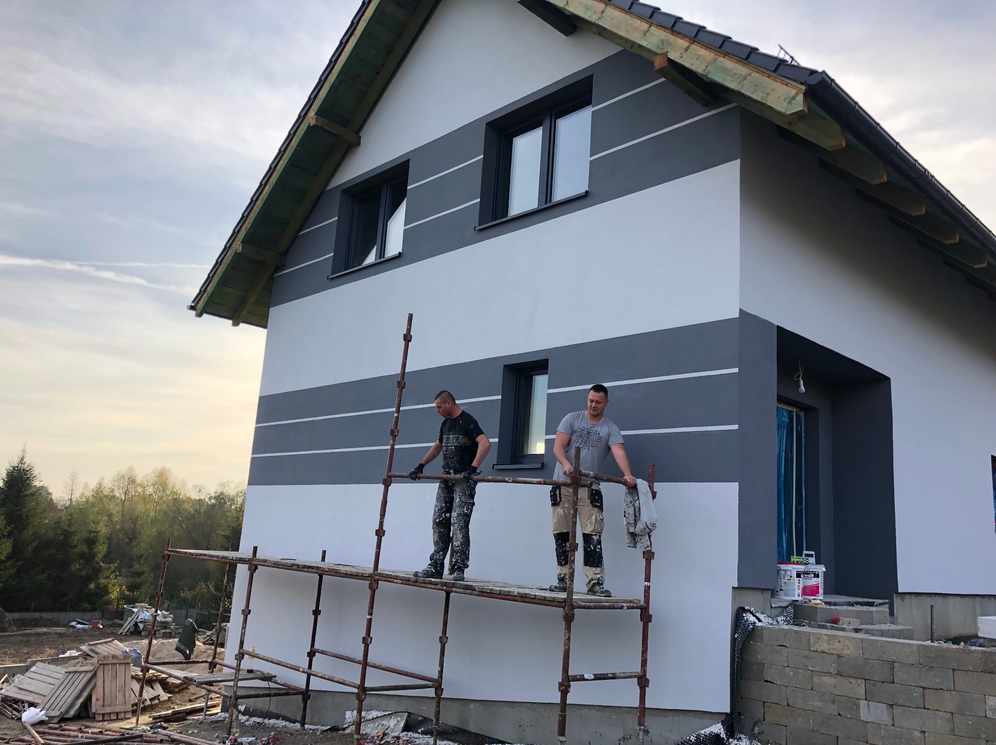 Dwóch mężczyzn na rusztowaniu maluje elewację domu w poziome pasy, szare i białe, z widocznymi oknami i fragmentem dachu.