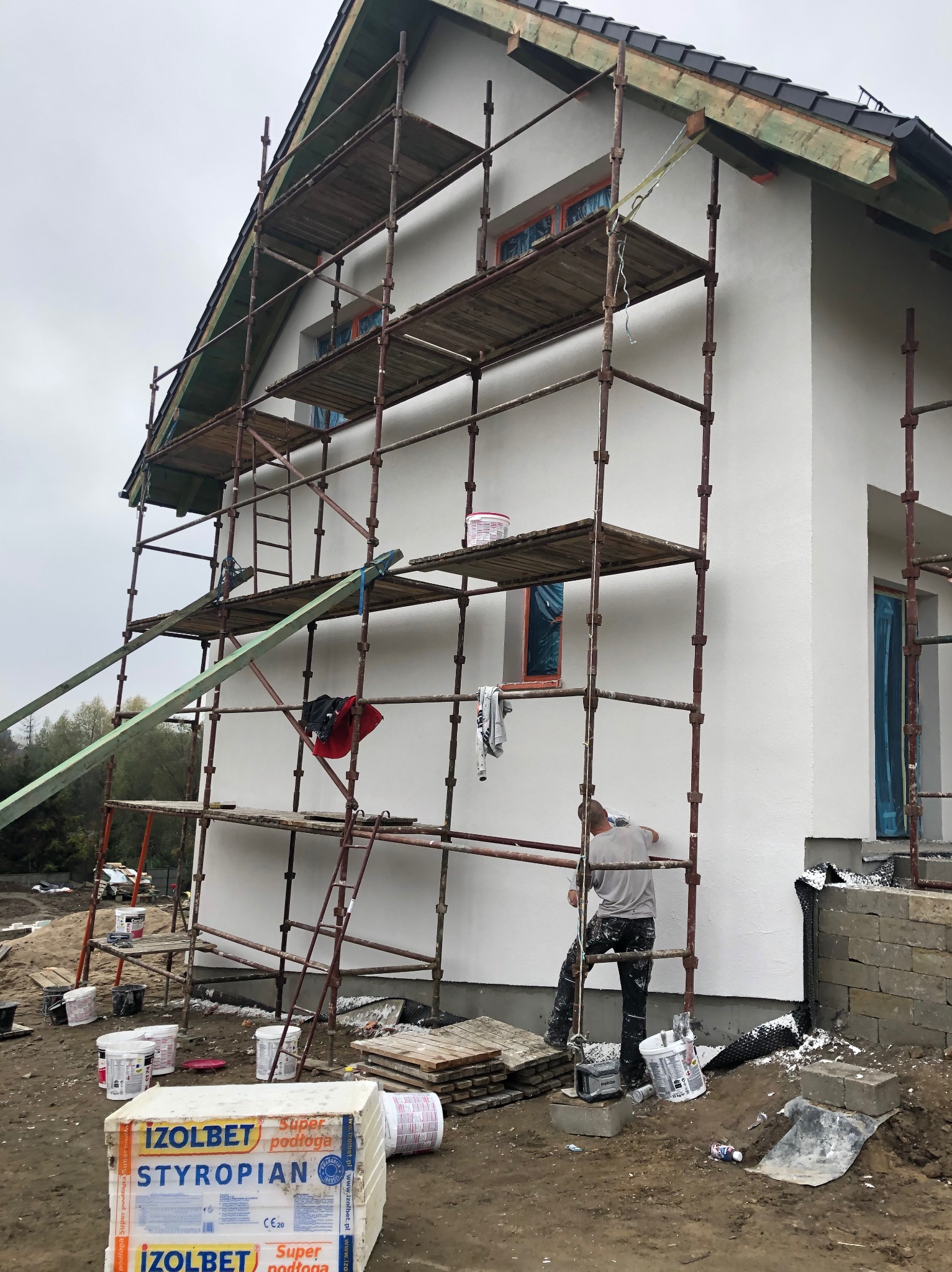 Pracownik na rusztowaniu podczas nakładania białego tynku na elewację domu jednorodzinnego, widoczne puszki z farbą, styropian, okna zabezpieczone folią.
