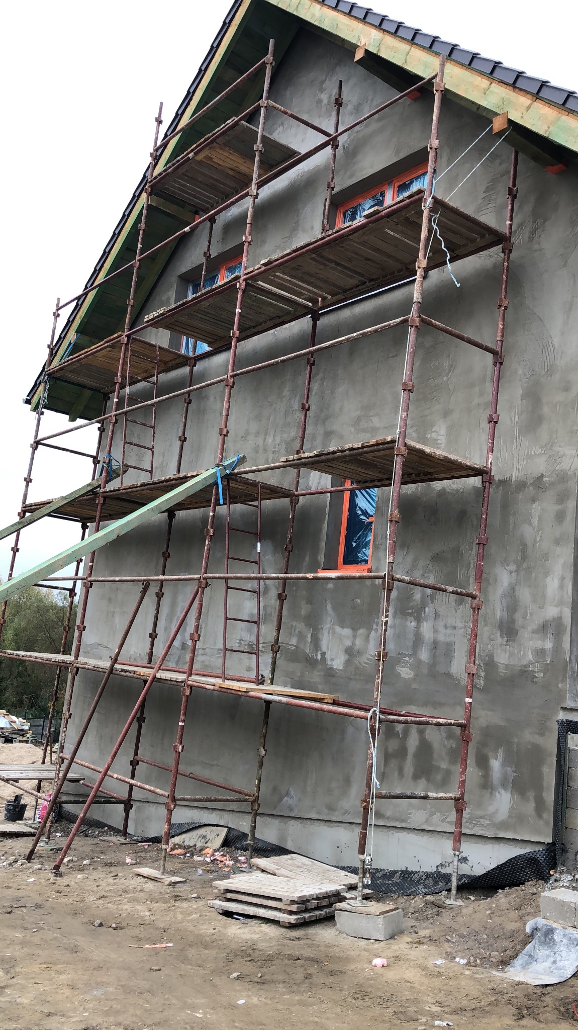 Budynek w trakcie nakładania szarego tynku, z zamontowanym rusztowaniem i zabezpieczonymi oknami folią ochronną.