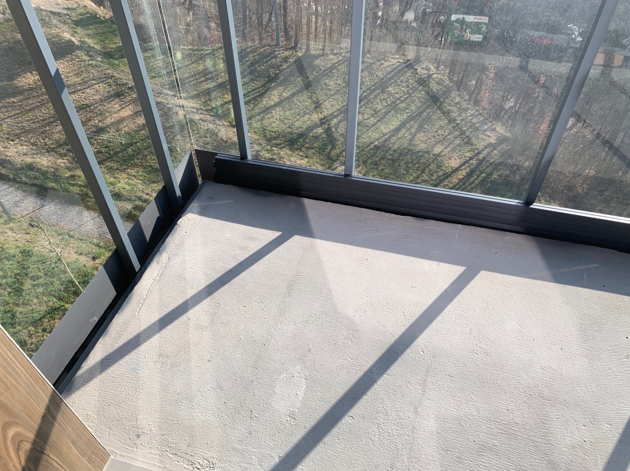 Szklana zabudowa balkonu z widokiem na drzewa, szare ramy okienne i betonowa podłoga w jasnym świetle słonecznym.