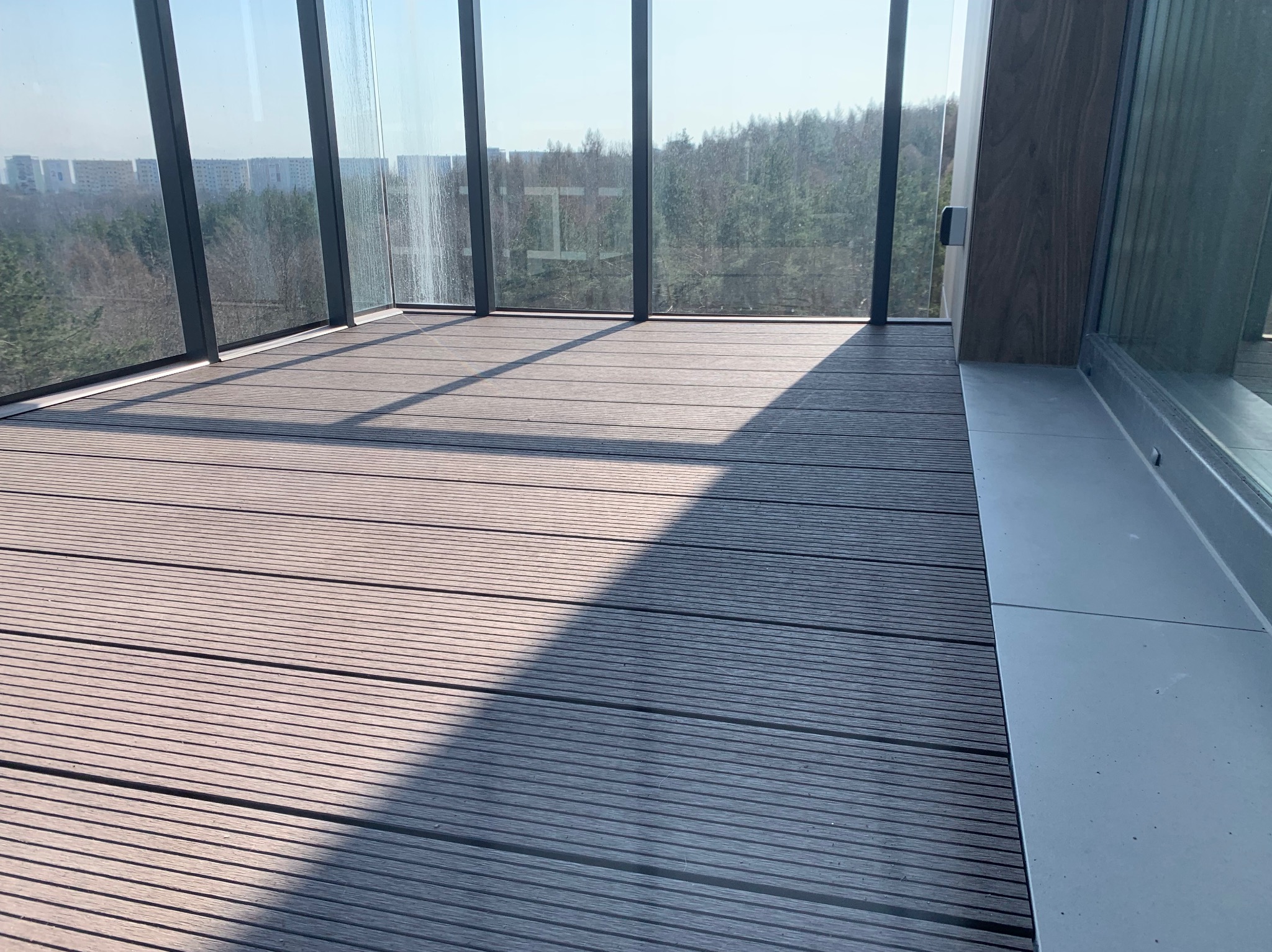 Nowoczesna zabudowa balkonu z drewnianą podłogą i panoramicznymi oknami z widokiem na las i zabudowę miejską w oddali.