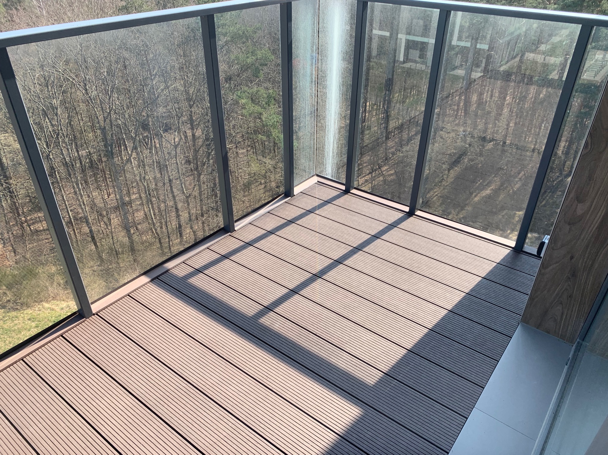 Nowoczesny balkon z szarą podłogą z desek kompozytowych i szklaną balustradą, widok na las w tle.