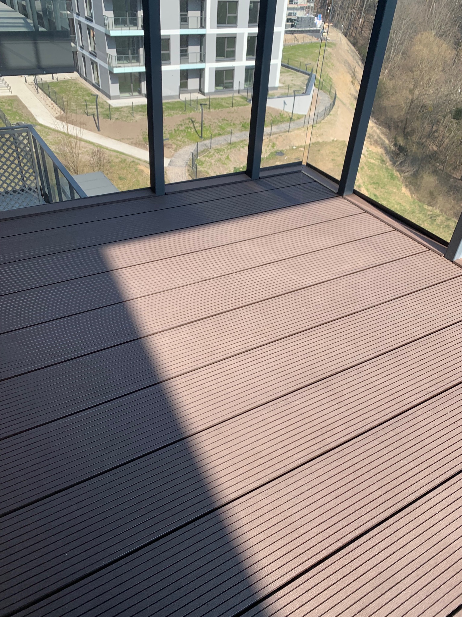 Nowoczesny balkon z podłogą z desek kompozytowych w odcieniach brązu, widok na osiedle mieszkaniowe i drzewa, fragment szklanej balustrady.