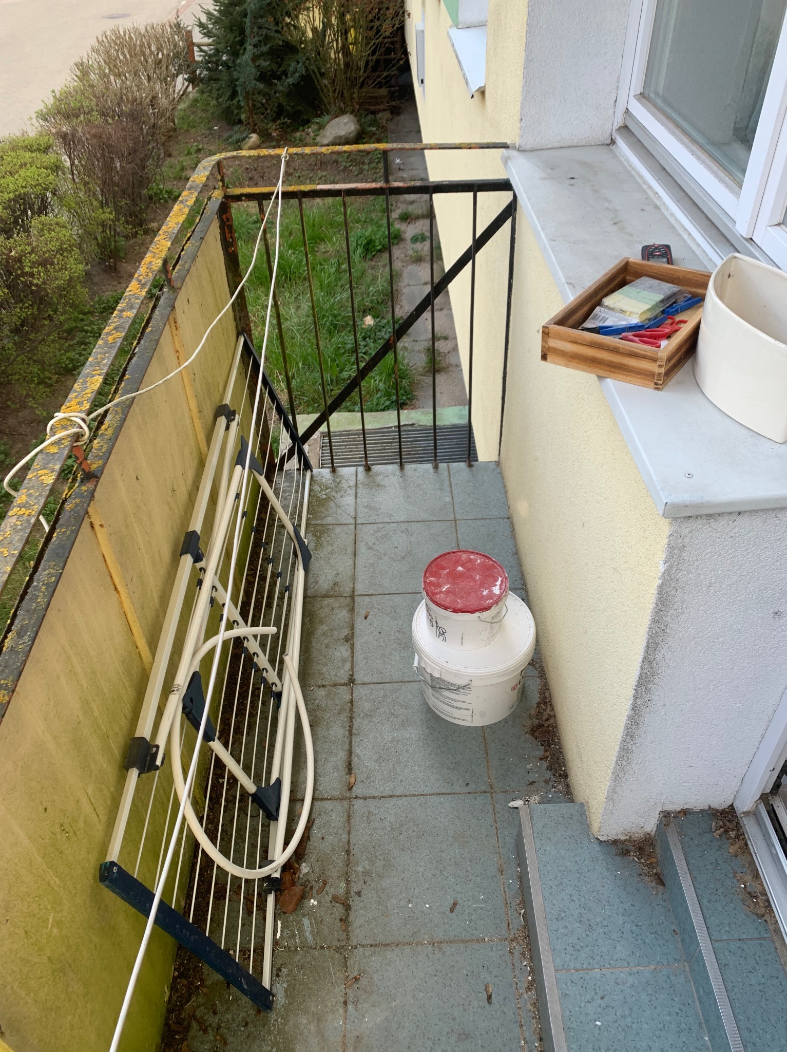 Przygotowania do odświeżenia balkonu w Gdańsku: widoczne wiadro z farbą z czerwoną pokrywką, paleta kolorów w drewnianej skrzynce oraz rozłożona suszarka na balkonie z metalową balustradą.