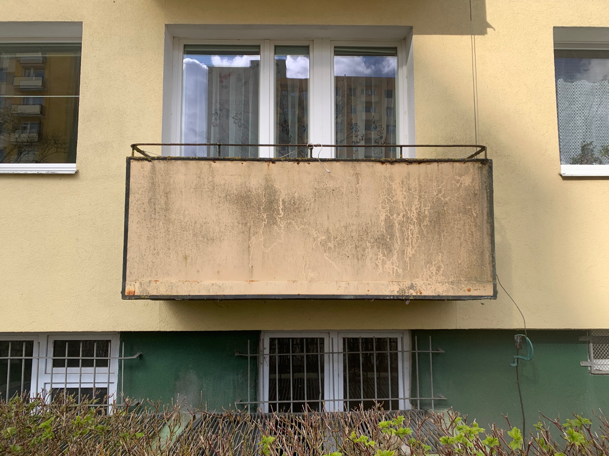 Balkon w bloku, z widocznymi śladami wilgoci i zabrudzeń na betonowej płycie, z metalową balustradą. W tle okna z firankami i fragment elewacji w odcieniach żółci i zieleni.