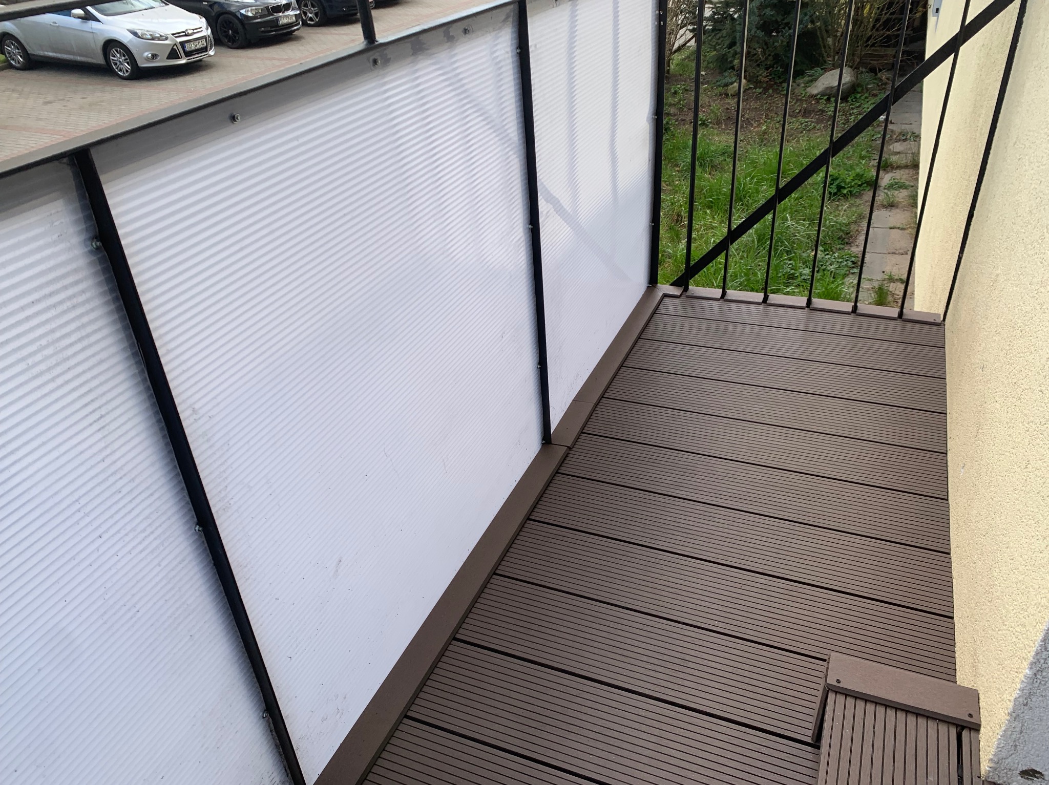 Zabudowany balkon z brązową podłogą z desek kompozytowych i ścianką z białego, prążkowanego tworzywa sztucznego z czarną ramą, widok z góry na fragment balkonu i otoczenie.