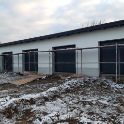 Letyro Bartosz Lekki - Biały budynek garażowy z czterema ciemnymi bramami segmentowymi, częściowo osłonięty rusztowaniem, na zewnątrz błoto i śnieg.