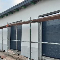 Letyro Bartosz Lekki - Montaż szarej segmentowej bramy garażowej z poziomo ułożonymi panelami, widok z rusztowania, w tle ocieplony budynek z szarą podbitką dachową, zimowy krajobraz.