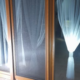 Letyro Bartosz Lekki - Okna przesuwne z moskitierą i firankami w drewnianej ramie, widok z zewnątrz na taras wyłożony płytkami.