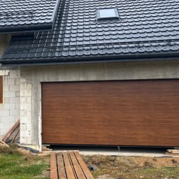 Letyro Bartosz Lekki - Zamknięta brama garażowa segmentowa w kolorze drewna, zamontowana w surowym budynku z białego bloczka, widoczne drewniane deski przed bramą.