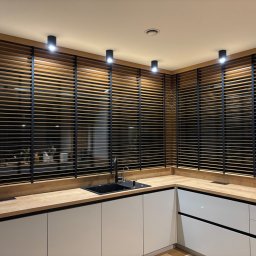 Letyro Bartosz Lekki - Drewniane żaluzje poziome w kolorze naturalnego drewna z czarnymi elementami, zamontowane w oknach nowoczesnej kuchni z białymi szafkami i drewnianym blatem.