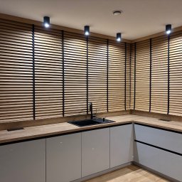 Letyro Bartosz Lekki - Jasne, drewniane żaluzje poziome z czarnymi taśmami w nowoczesnej kuchni z drewnianym blatem i szarymi szafkami, oświetlone punktowymi lampami sufitowymi.