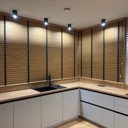 Letyro Bartosz Lekki - Drewniane żaluzje poziome z czarnymi taśmami w nowoczesnej kuchni z białymi szafkami i drewnianym blatem, oświetlone punktowymi lampami sufitowymi.