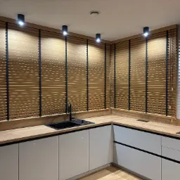 Drewniane żaluzje poziome z czarnymi taśmami w nowoczesnej kuchni z białymi szafkami i drewnianym blatem, oświetlone punktowymi lampami sufitowymi.