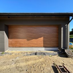 Letyro Bartosz Lekki - Nowa, segmentowa brama garażowa z drewnopodobnym panelem, zamontowana w nowoczesnym domu z szarą elewacją, widoczna w trakcie prac wykończeniowych na zewnątrz.