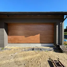 Nowa, segmentowa brama garażowa z drewnopodobnym panelem, zamontowana w nowoczesnym domu z szarą elewacją, widoczna w trakcie prac wykończeniowych na zewnątrz.